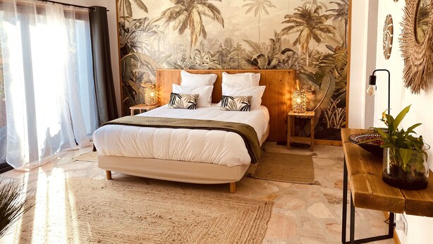 A bedroom with a large bed, a wooden headboard, a nightstand with a lamp, a potted plant, and a tropical mural on the wall. - Le Mas de San lou (Cabasse)