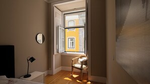 Un dormitorio con una cama, una silla, un escritorio, una lámpara y una ventana con vistas a edificios.