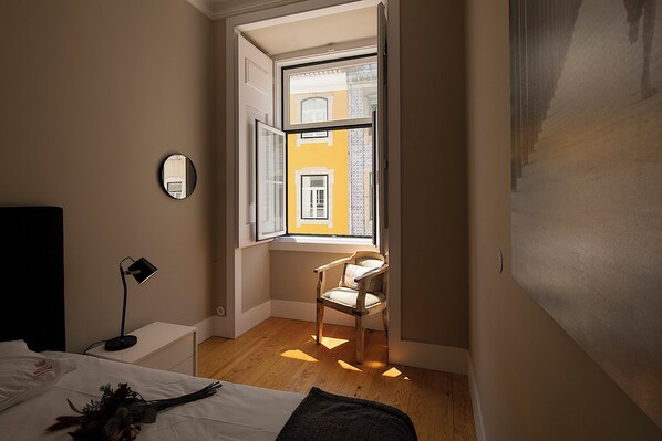 Un dormitorio con una cama, una silla, un escritorio, una lámpara y una ventana con vistas a edificios.