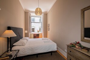 Un dormitorio con cama, mesita de noche, lámpara, cómoda, ventana y espejo.