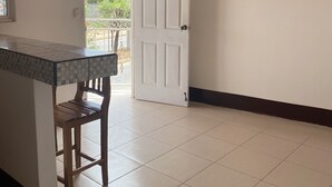 Una habitación con suelo de baldosas, techo de madera, una puerta blanca, una pequeña mesa con una silla y un cuadro enmarcado en la pared.
