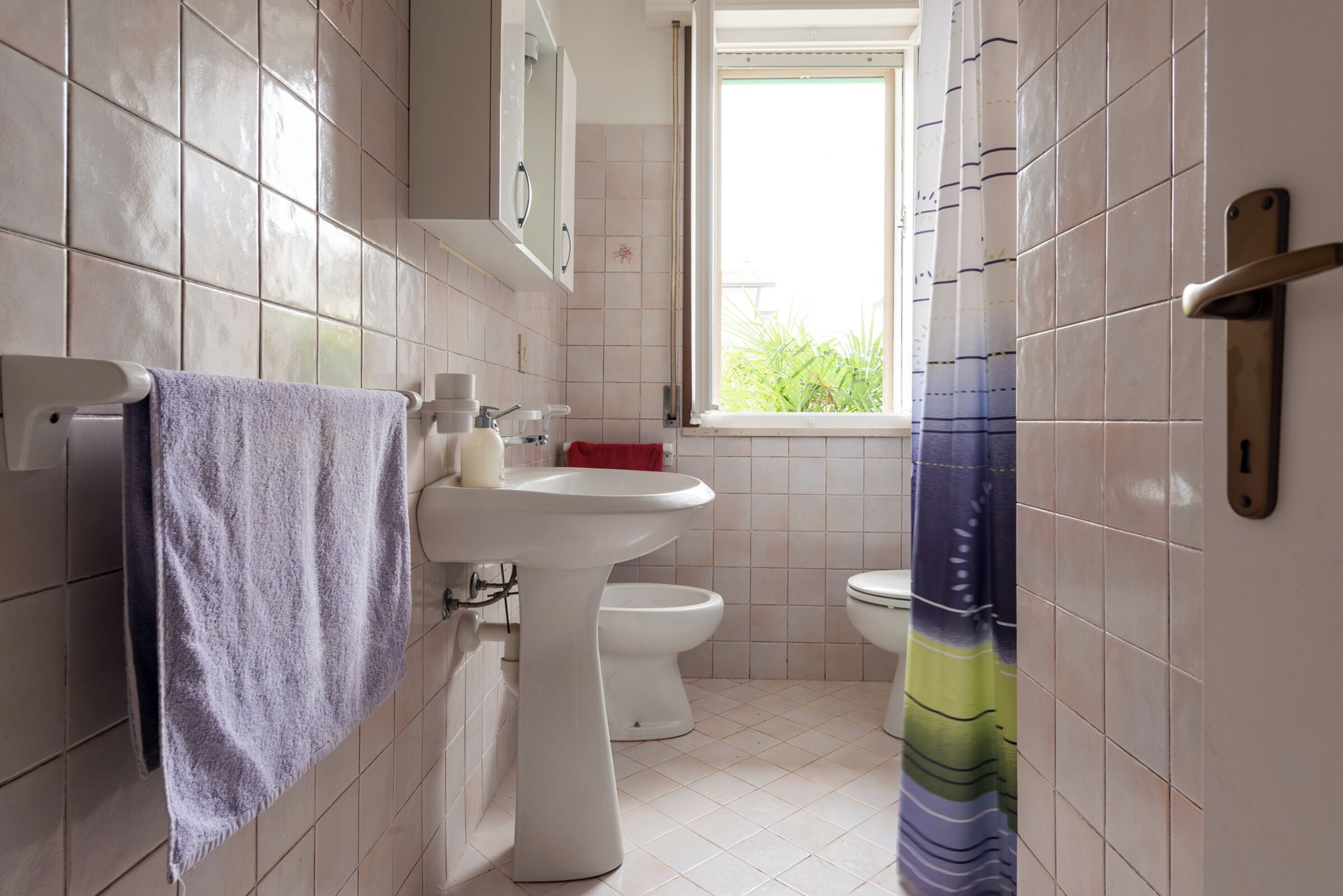 A bathroom with a white pedestal sink, a white toilet, and a window with a view of greenery.