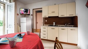 A compact kitchen with a dining area, featuring a table with a red polka dot tablecloth, a television mounted above the counter, and a refrigerator to the left. - Luminous Apartment With Garden (San Michele al Tagliamento)