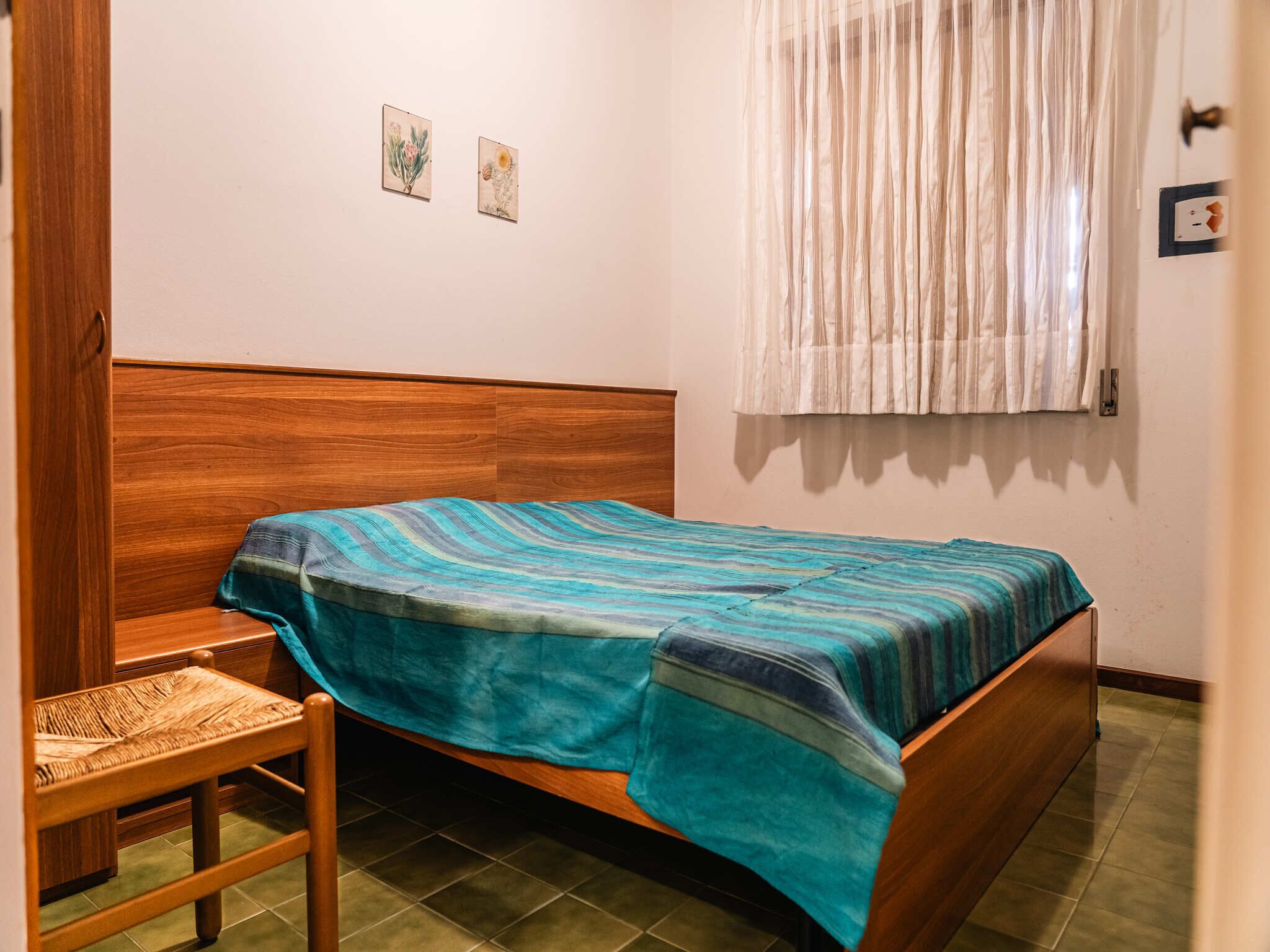 A room with a wooden bed, a small wooden chair, a window with sheer curtains, and two wall-mounted pictures.