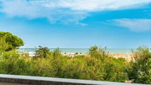 A beach view with a clear sky, trees in the foreground, and a distant beach area with umbrellas. - Beautiful Flat With sea View in Bibione (San Michele al Tagliamento)