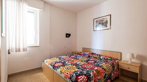 A bedroom with a bed, a nightstand, a lamp, a window with curtains, and a picture on the wall. - Bright Flat Just Steps From Bibione Beach (San Michele al Tagliamento)