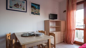 A small, well-lit room with a wooden table set for dining, two chairs, a television on a shelf, and a large window with curtains. - Relax in our Flat With sea View Terrace (San Michele al Tagliamento)