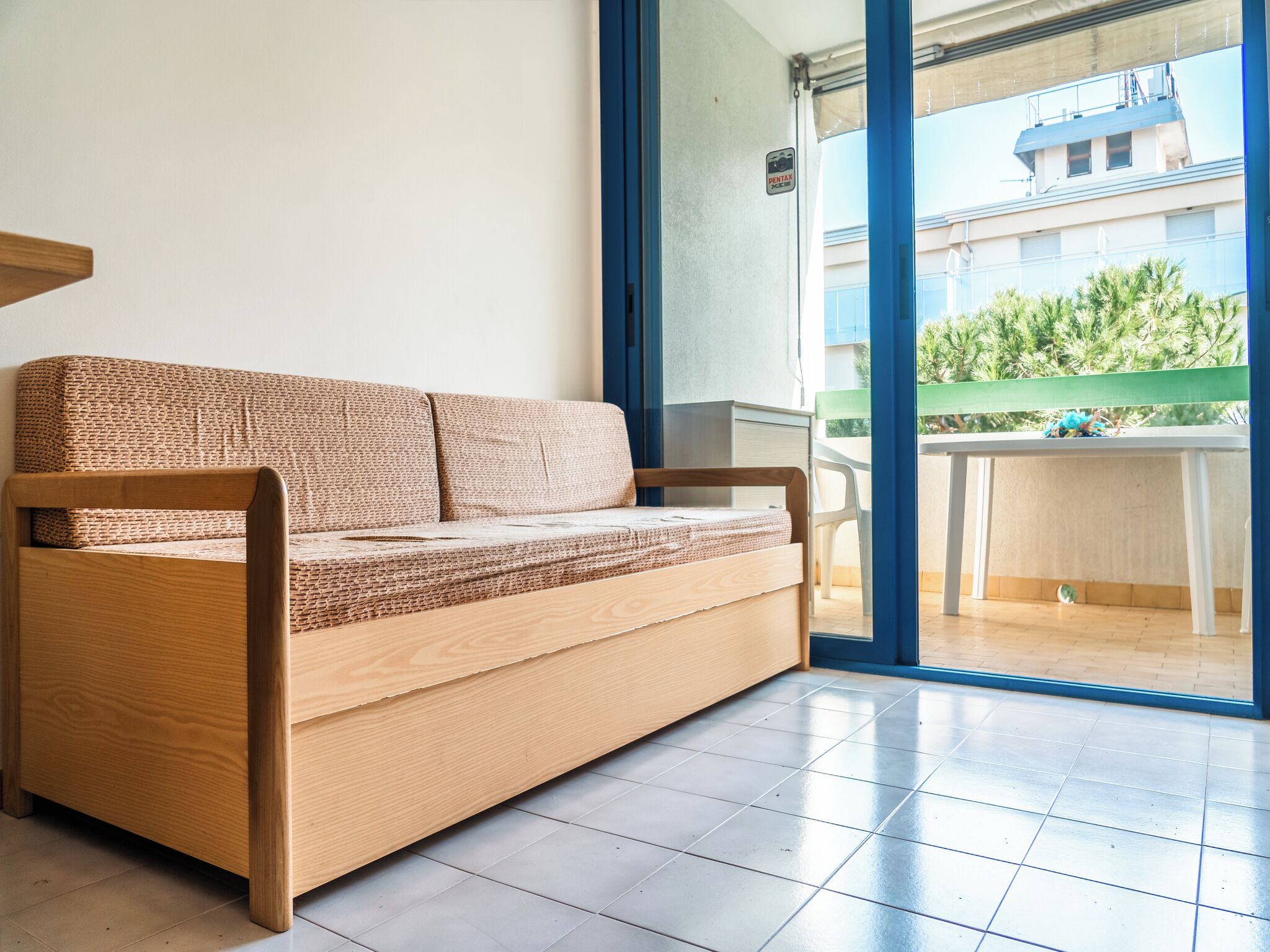 A wooden sofa with a woven cover in a room with a tiled floor and a sliding glass door leading to an outdoor balcony.