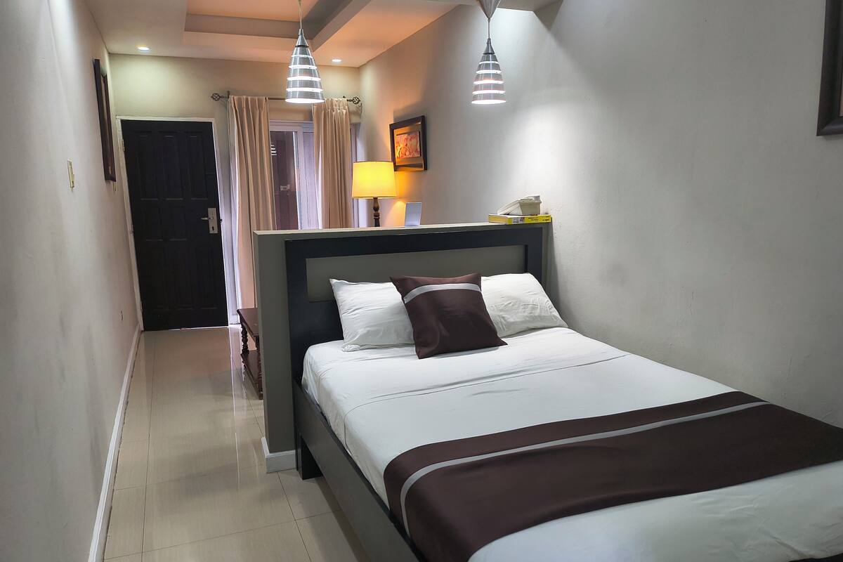 Une chambre d’hôtel avec un lit, une table de chevet, une lampe, un tableau au mur et une porte donnant sur une autre pièce.