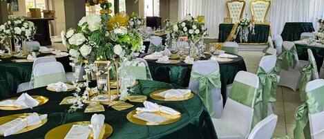 Un salón de banquetes con mesas redondas cubiertas con manteles verdes, sillas blancas con cintas verdes y centros de mesa florales.