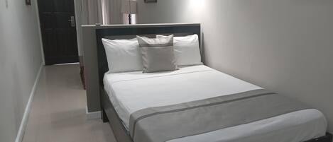 A neatly made bed with white linens and a grey accent, a bedside lamp, and a framed picture on the wall.