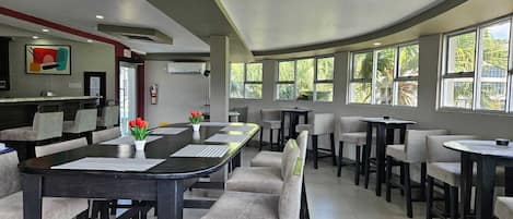 Un comedor moderno con una mesa larga y múltiples sillas, amplias ventanas con vistas a la vegetación, y una cocina visible al fondo.