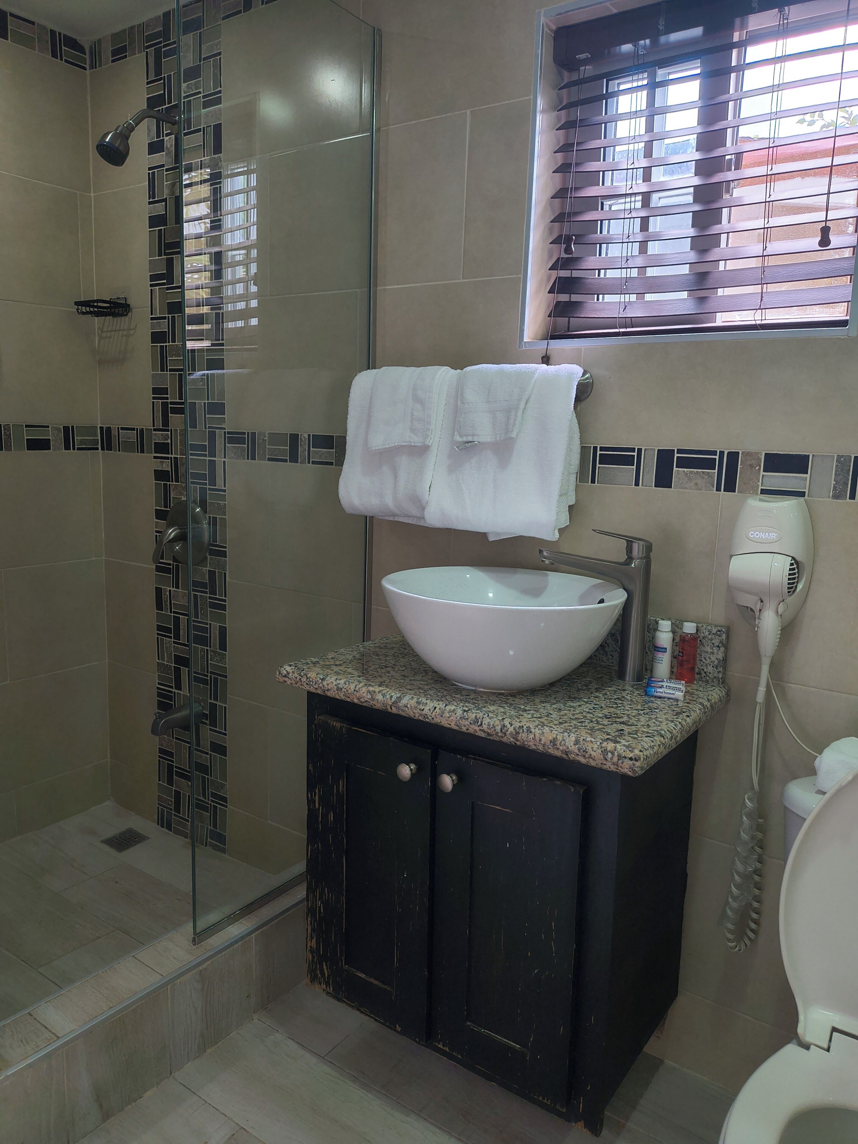 Un baño con ducha envuelta en vidrio, un lavamanos blanco con lavabo, un mueble de tocador con un armario oscuro y una ventana con persianas.