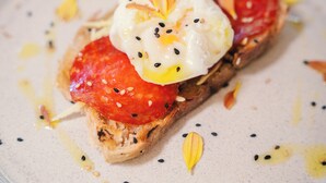 Ein Teller mit einem Sandwich, belegt mit Tomate, Ei und Saaten, dazu eine kleine Schale Sauce.
