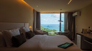 Ein Hotelzimmer mit einem großen Bett, einem Schreibtisch, einem Sessel und Blick auf das Meer.