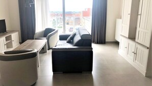A modern living room with a sofa, a round ottoman, a TV on a cabinet, and a view of buildings through large windows.