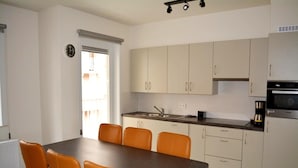 A modern kitchen with white cabinets, a dining table with four orange chairs, a built-in oven, and a clock on the wall.