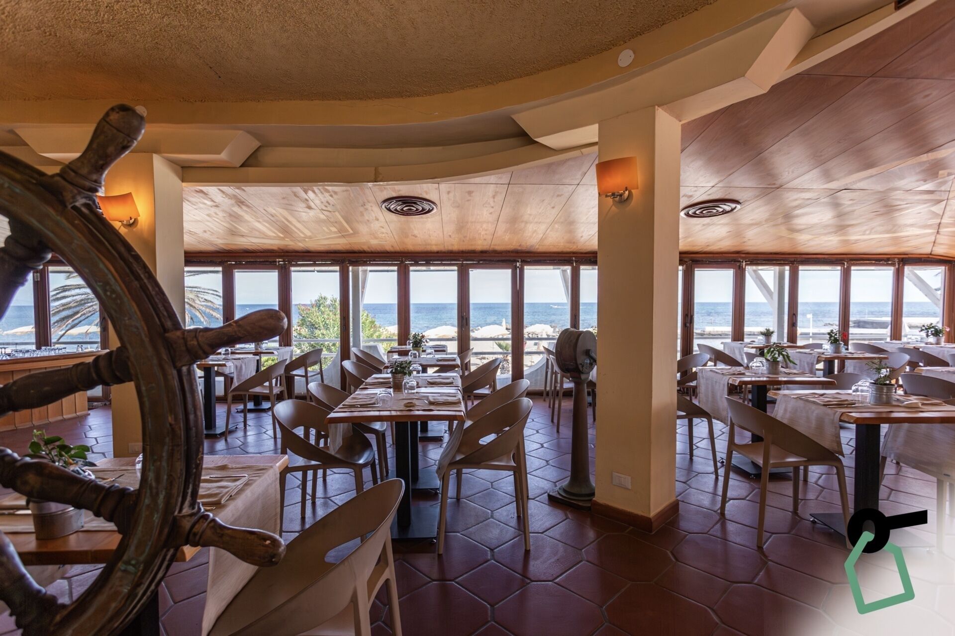 Un restaurant doté d’un sol carrelé à motifs hexagonaux, de tables et de chaises en bois, et de grandes fenêtres offrant une vue sur la mer.