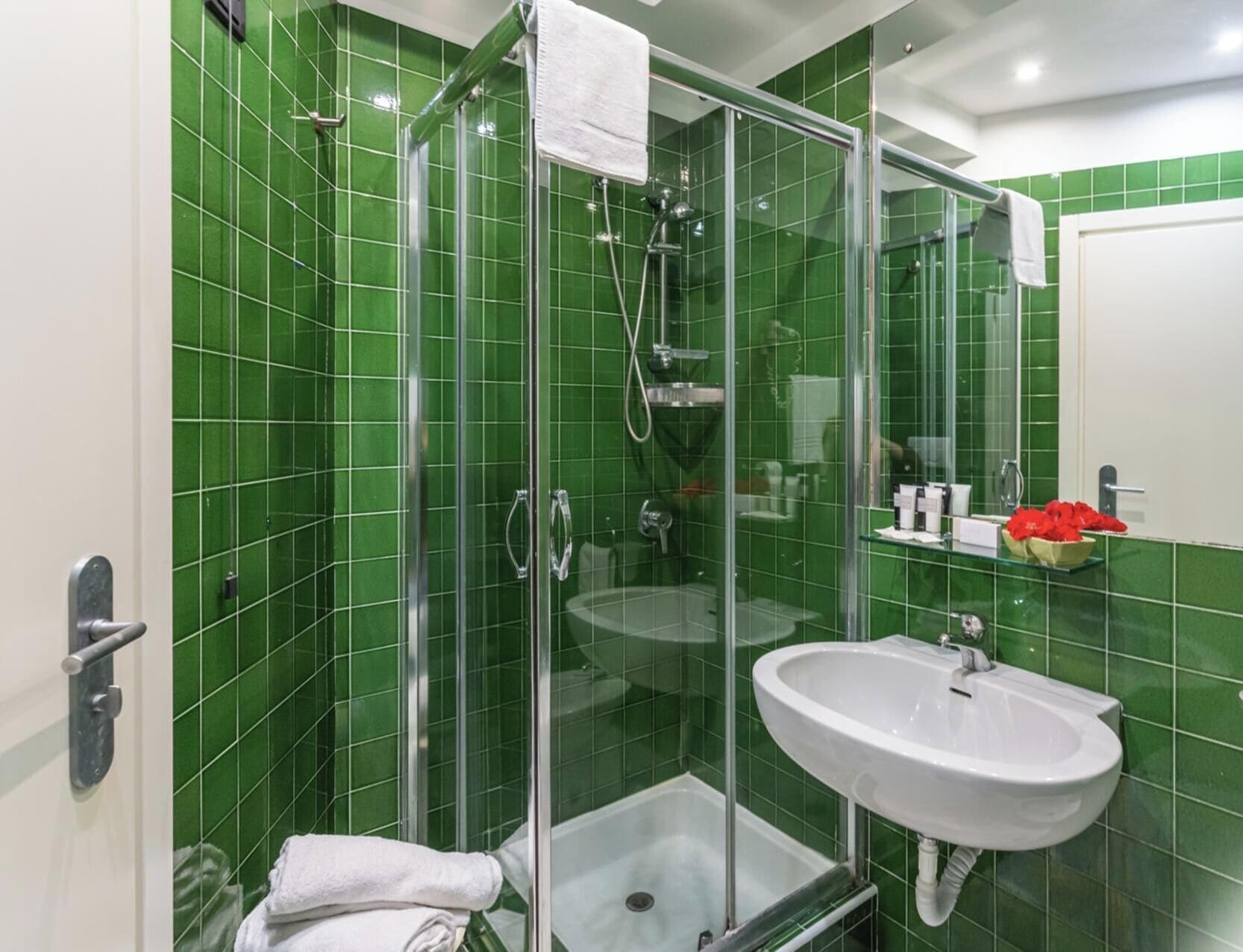 A bathroom with green tiles, a glass shower enclosure, a white sink, and a towel rack.