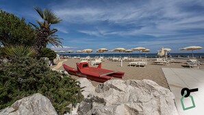 A beach with rows of sun loungers and umbrellas, a red boat, and rocky terrain. - Hotiday Room Collection - Circeo (San Felice Circeo)