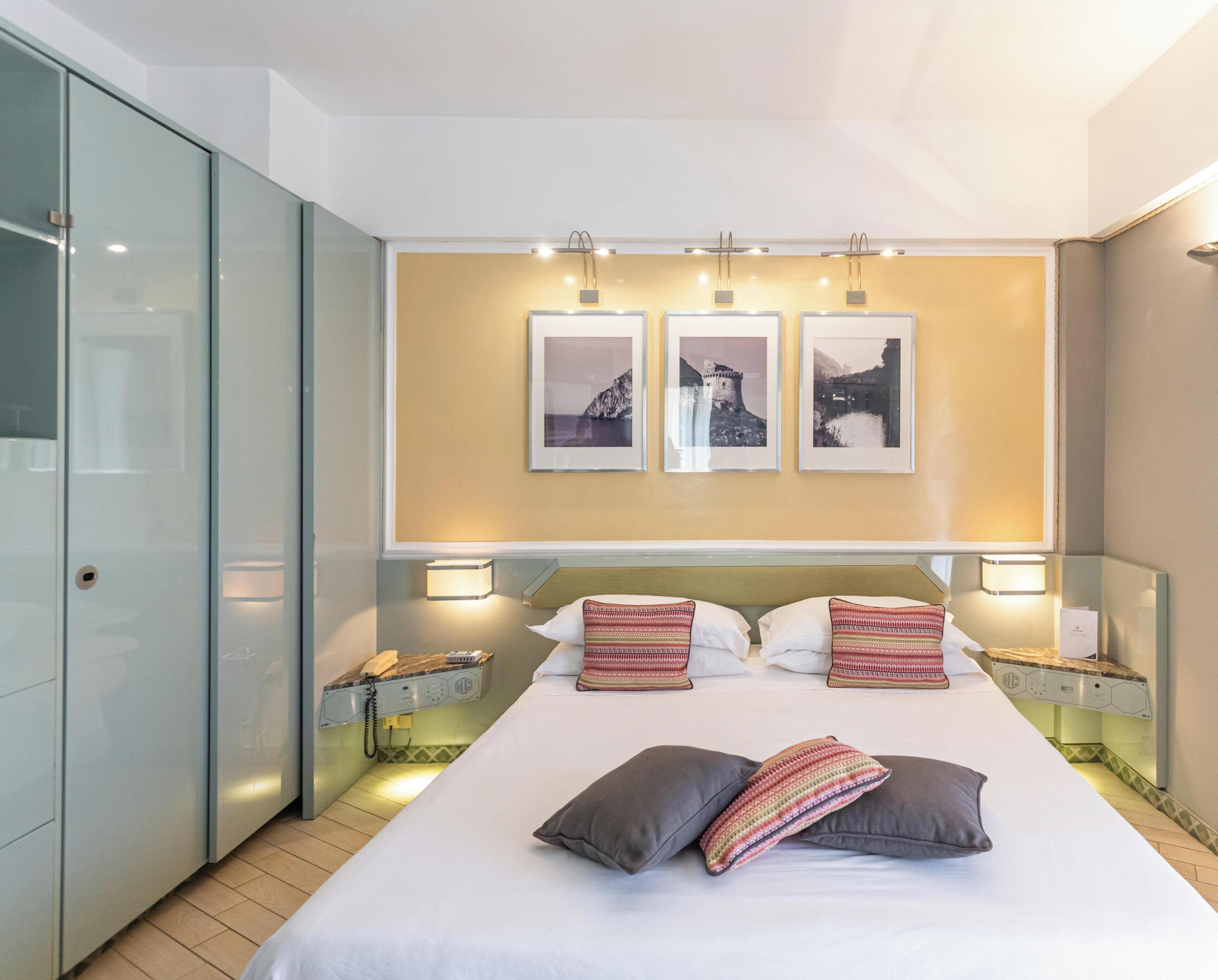 Une chambre d’hôtel avec un lit, deux tables de chevet, un téléphone et trois tableaux encadrés accrochés au mur.