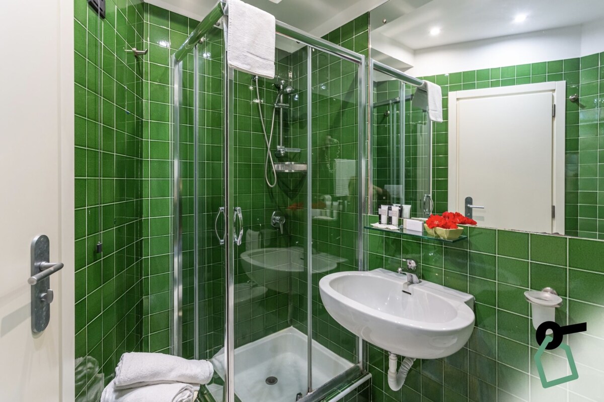 A bathroom with green tiles, a glass-enclosed shower, a sink, and a white door.