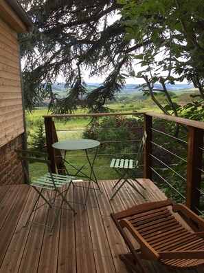 Una terraza de madera con una mesa y sillas, con vistas a un paisaje verde con árboles y colinas al fondo.