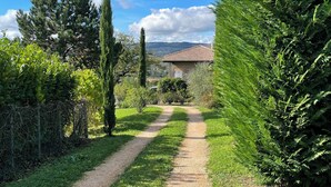 Un chemin de gravier mène à une maison dont le toit est recouvert de tuiles, et qui est entourée d’une végétation luxuriante et d’arbres.