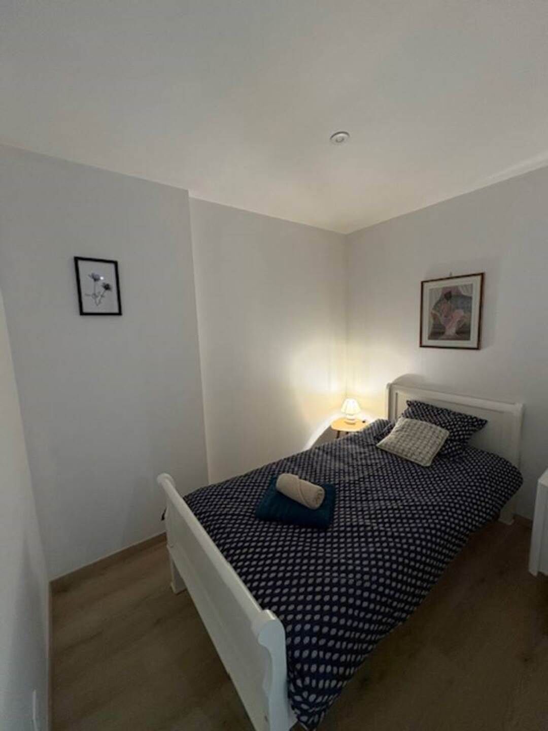 A small, neatly arranged bedroom with a single bed, a bedside table, a lamp, and two framed pictures on the walls.