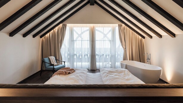 A bedroom with a sloped ceiling, a large bed, a chair, a bathtub, and a view through a window with curtains.