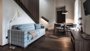 A modern living room with a blue plaid sofa, a dining table with chairs, a floor lamp, and a TV mounted on the wall.