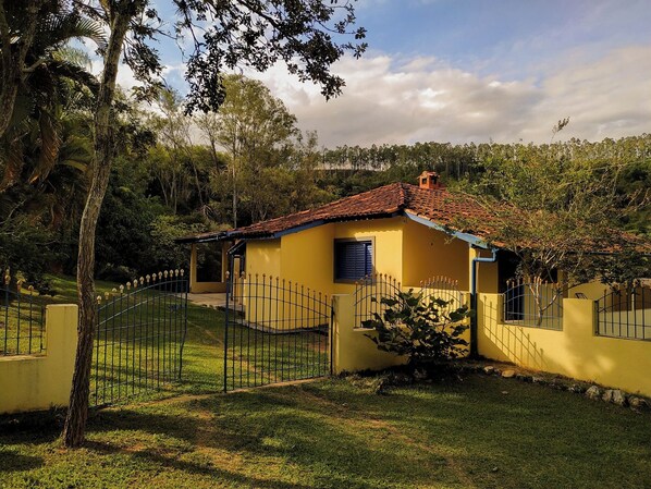 Una casa amarilla con techo de tejas rojas, rodeada por una cerca de metal y árboles.