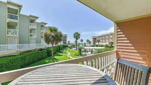 A balcony with a view of a well-maintained garden, a wooden deck, and a building with multiple balconies and a pool area.