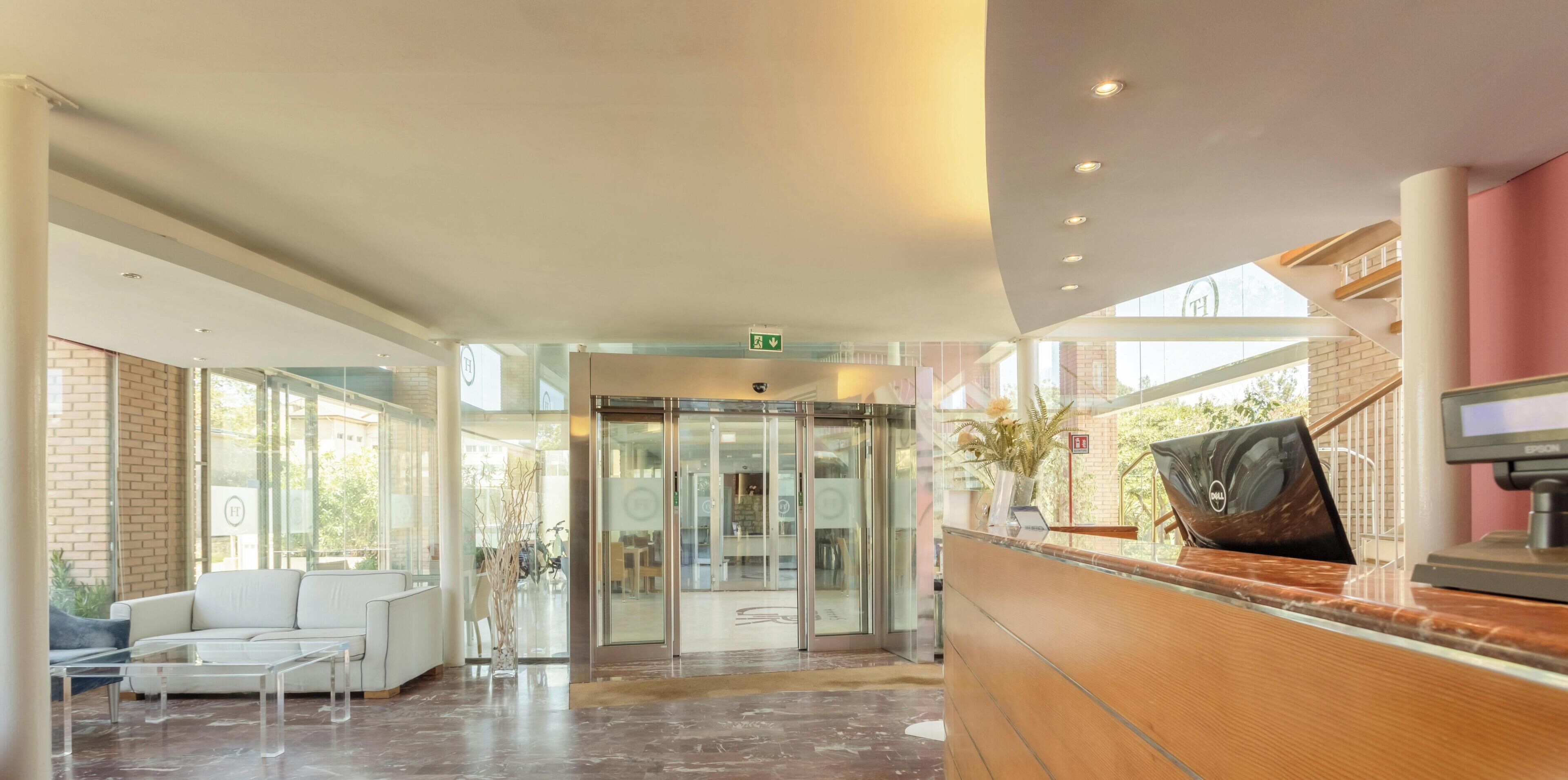 Un espace d’accueil moderne doté d’un ascenseur en verre, d’un comptoir de réception et d’un coin salon avec un canapé blanc et une table basse.