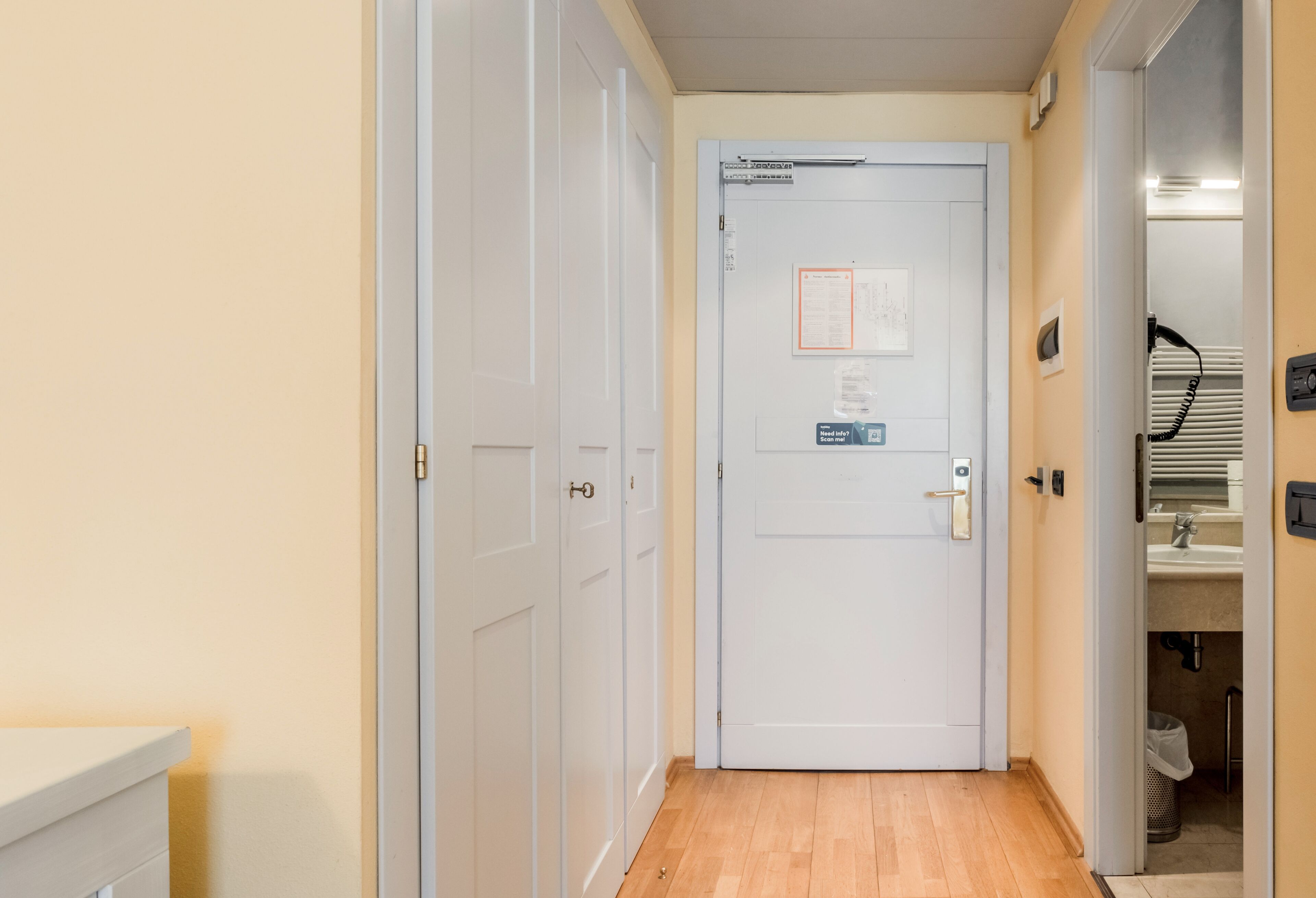 A narrow hallway with a white door, a small sign, and a visible trash bin.