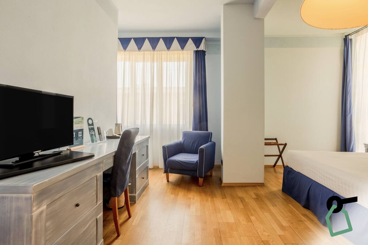 A hotel room with a flat-screen TV, a desk, a chair, a bed, and a window with curtains.