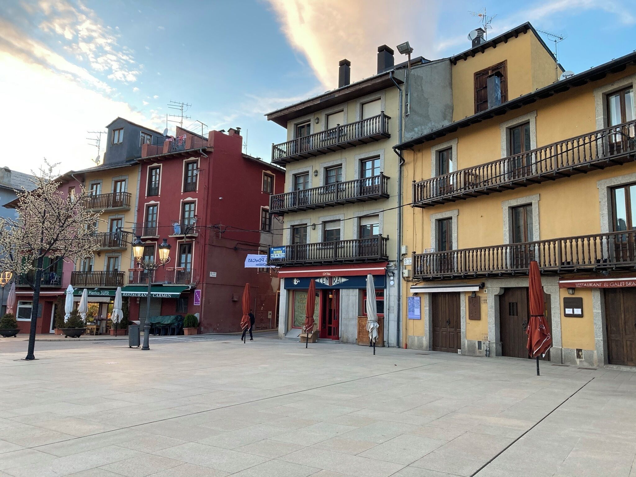 Une place bordée de bâtiments, avec des terrasses et un ciel dégagé.