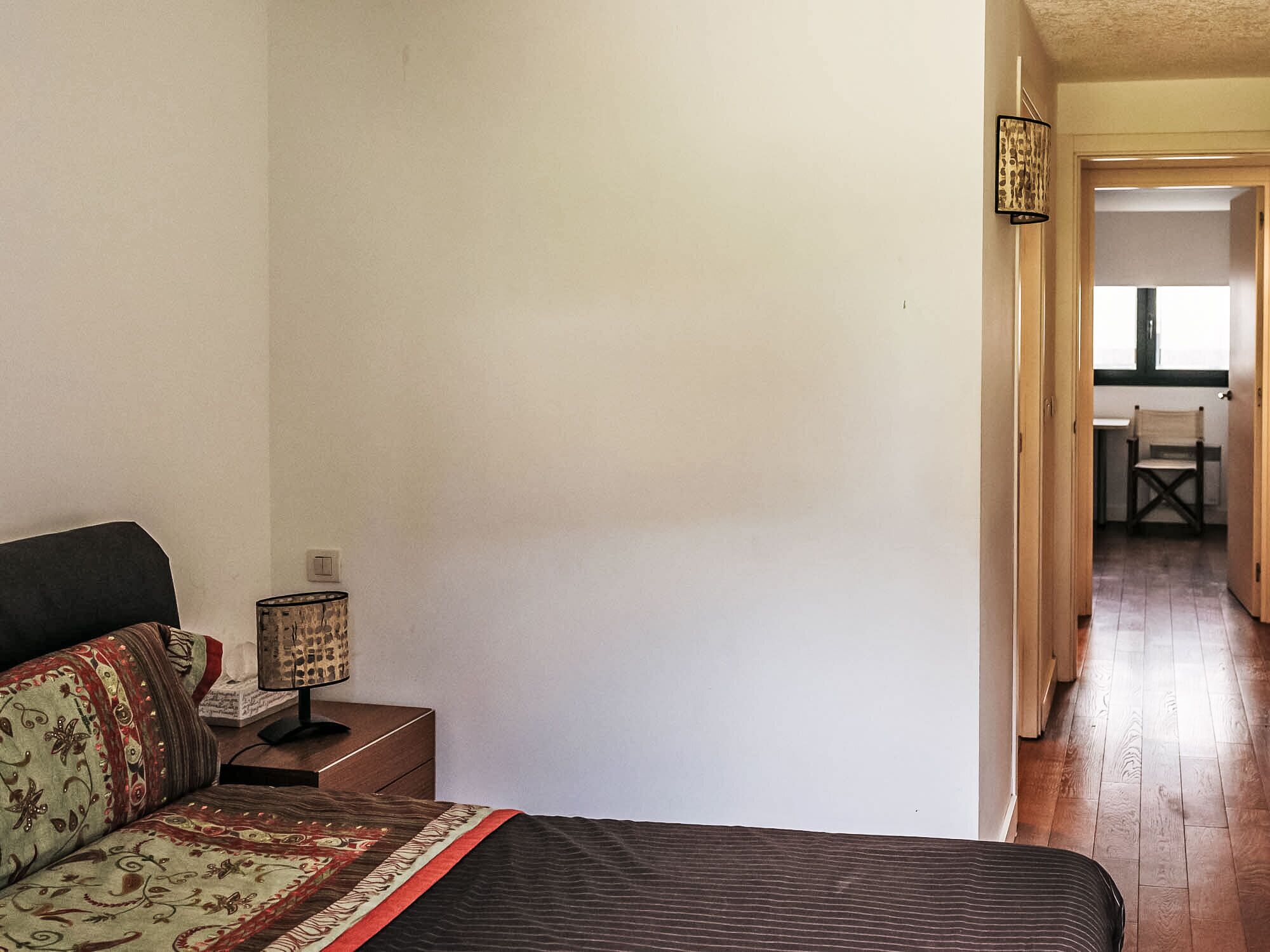 A bedroom with a bed, a nightstand, a lamp, and a chair in the background.