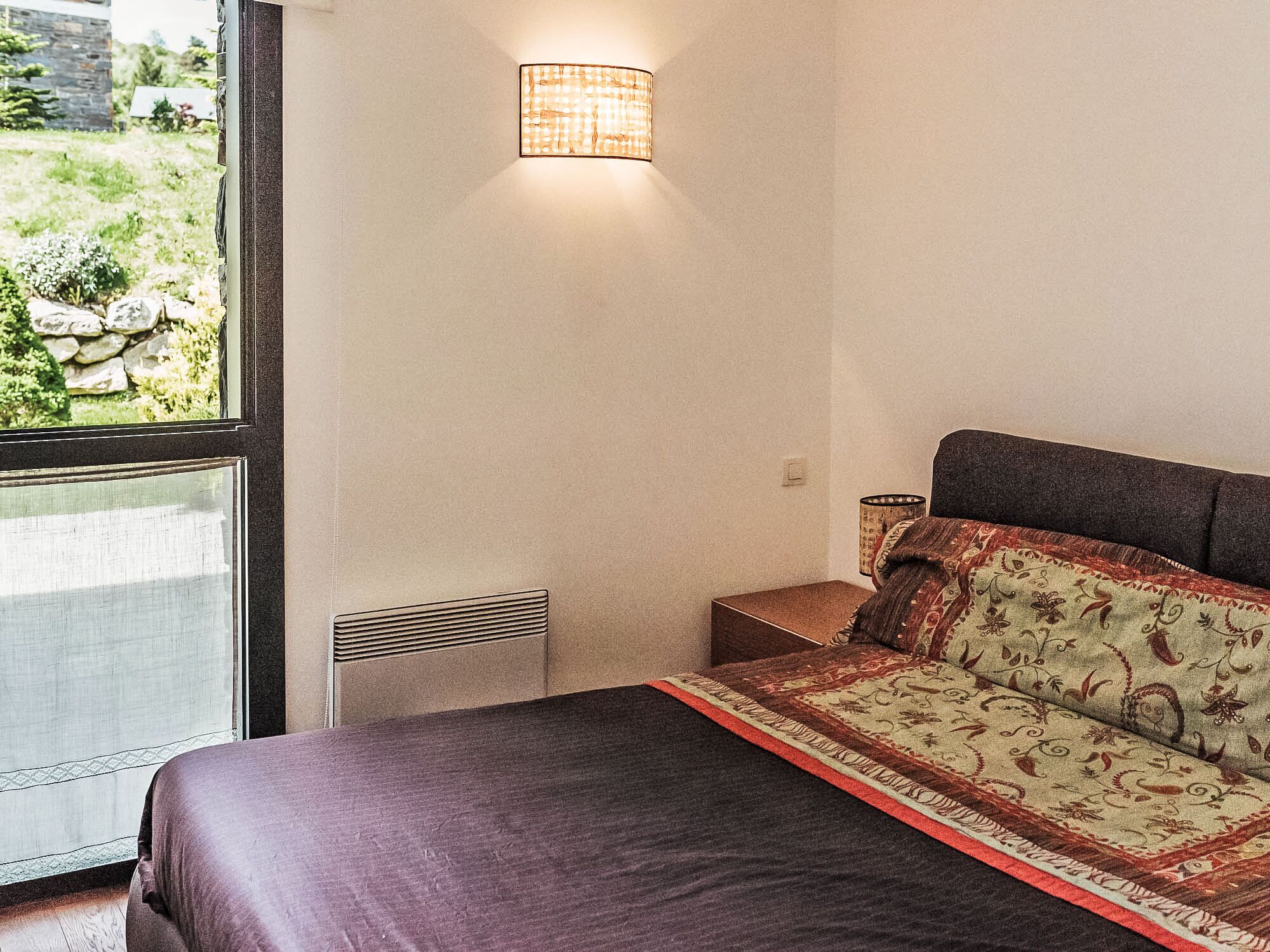 A bedroom with a large window, a bed with a patterned bedspread, a nightstand, and a wall-mounted lamp.