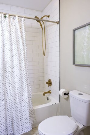 A bathroom with a white tiled shower, a gold showerhead, a white toilet, and a framed picture on the wall. - The Bluegrass Railway Inn (Midway)