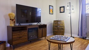 A living room with a flat-screen TV on a wooden cabinet, a round coffee table with a board game, a rustic barrel, and a floor lamp. - The Bluegrass Railway Inn (Midway)