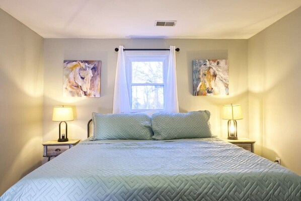 A bedroom with a bed, two bedside lamps, a window with a curtain, and two wall-mounted paintings. - The Bluegrass Railway Inn (Midway)