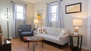 A living room with a grey sofa, a blue armchair, a round coffee table, and a small side table with a lamp. There are two windows with curtains, a framed picture on the wall, and a floor lamp. - The Bluegrass Railway Inn (Midway)