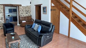 Uma sala de estar com um sofá de couro preto, uma mesa de centro de madeira e um tapete estampado. Uma escada com corrimãos de madeira leva a um andar superior.