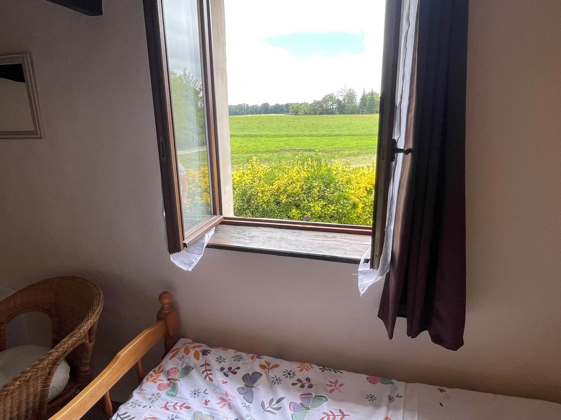 Un dormitori amb un llit individual, una finestra amb vista a un camp verd i una cadira de vimet.
