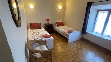 A small, cozy room with two single beds, a small table, a chair, and a window with curtains.