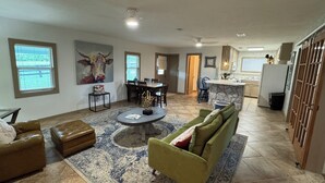 A spacious living room with a green sofa, a brown leather couch, and a dining area with a table and chairs. There is a kitchen area with a refrigerator and a window. - 3-Br Nature Oasis in rolling hills of Burton, 15 minutes from Round Top (Burton)