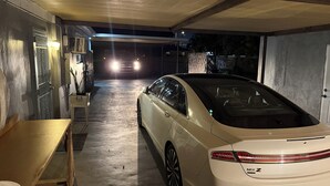 Une voiture blanche est garée dans un garage faiblement éclairé, dont le sol est en béton, avec un banc en bois et une porte métallique en arrière-plan.