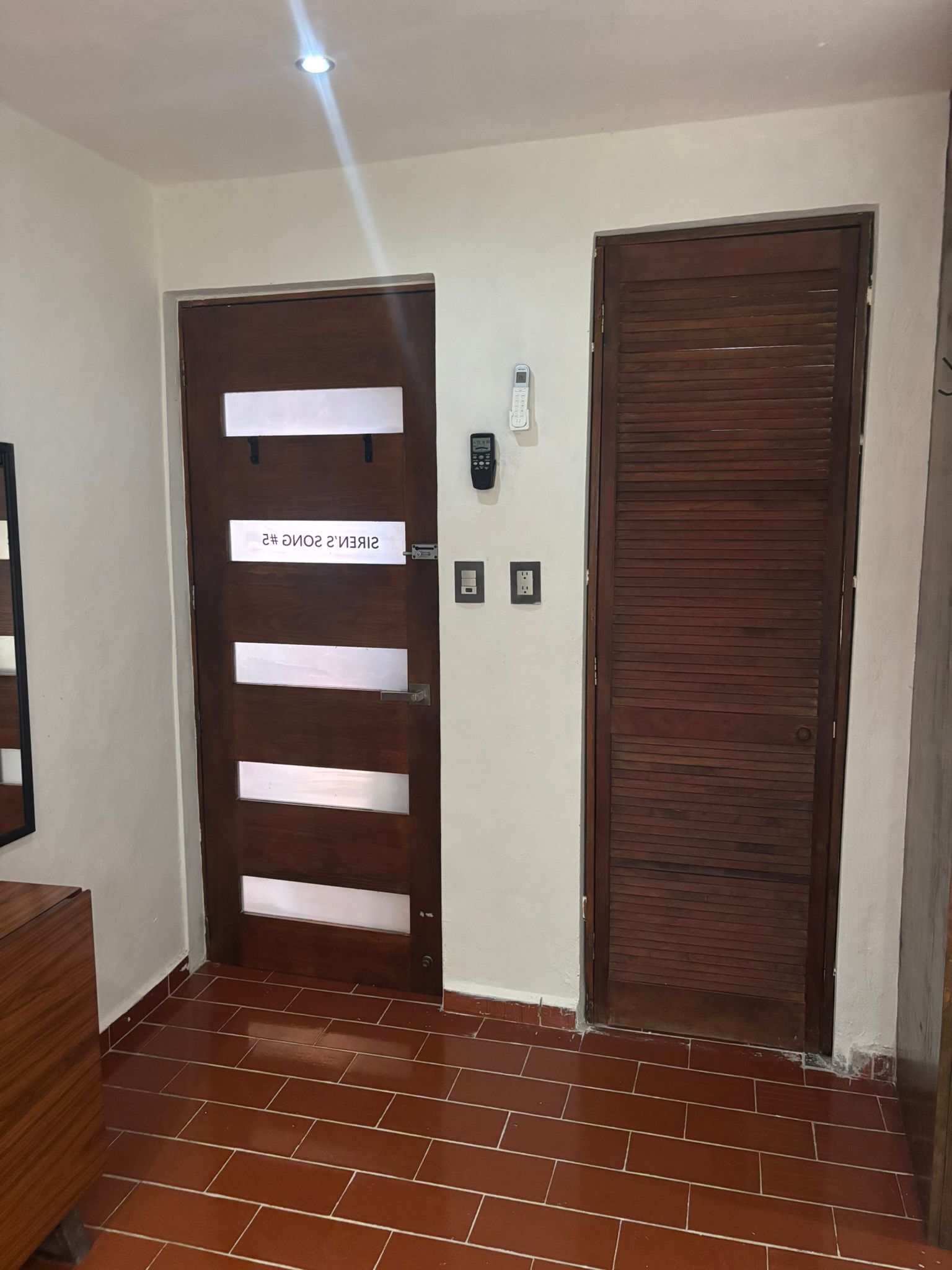 Un pasillo con una puerta de madera que dice 'SIREN'S 2' y otra puerta de madera cerrada. Hay dos enchufes eléctricos e interruptor de luz en la pared.