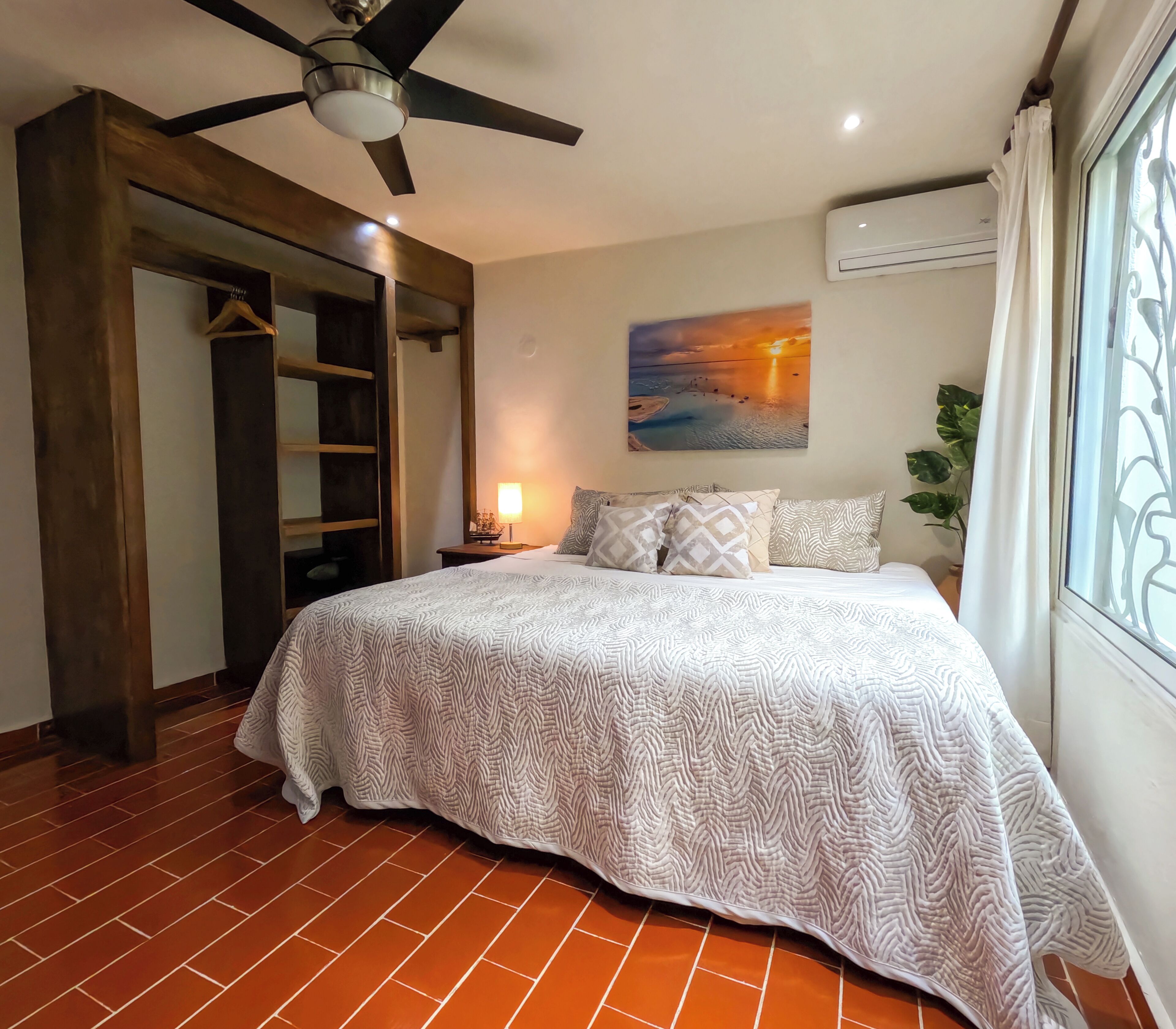 Une chambre à coucher avec un lit, un ventilateur de plafond, une table de chevet avec une lampe, un tableau au mur et une fenêtre avec des rideaux.